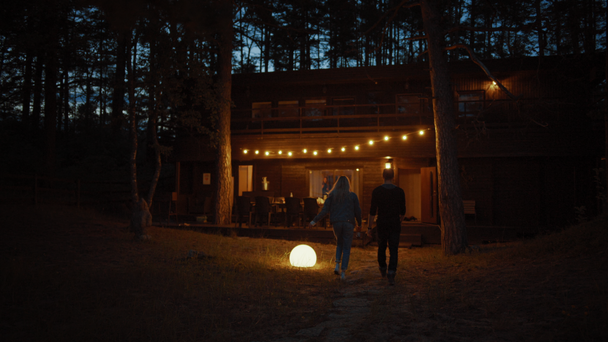 Genç Adam ve Kadın Birlikte Bir Cozy Cottage Walk. House'da Teras'ın Üzerinde Asılı Sıcak Işıklar Var. Romantik Yaz Akşamı Atmosferi. Ev Bir Çam Ormanı'nda yer almaktadır. - Video, Çekim