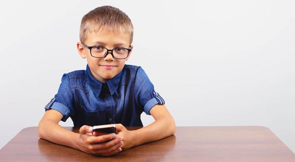 白い背景に学校の机の上に携帯電話を使用して眼鏡をかけた笑顔の少年のバナー肖像画 現代のモバイルと通信 - 写真・画像