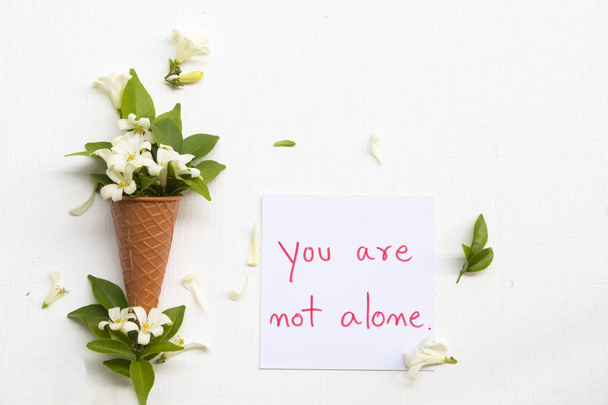 you are not alone message card handwriting with white flowers jasmine in cone arrangement flat lay postcard style on background white  - Фото, зображення