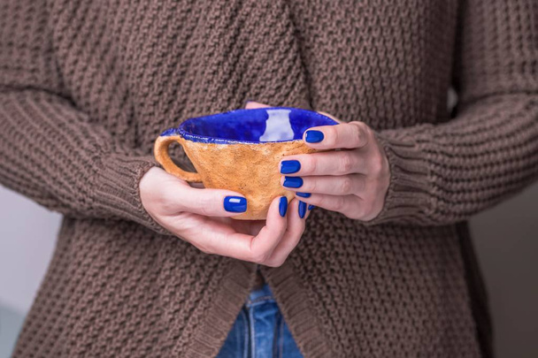 Krásné ženské ruce držící hnědou hliněnou šálku s modrou sklovkou. Manikúra s modrým leštěkem na nehty. Útulný koncept - Fotografie, Obrázek