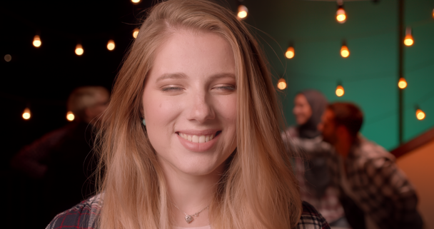 Closeup portrait of young pretty blonde long haired female looking at camera at diverse party in cozy evening - Footage, Video