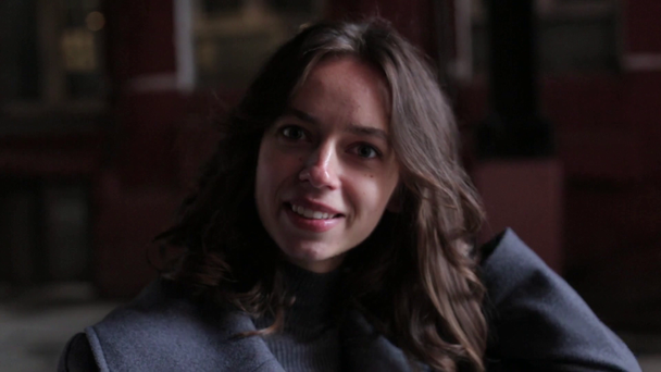 ein junges Mädchen in grauem Mantel und Rollkragen mit Armbanduhr auf der Hand blickt in die Kamera, richtet ihr welliges Haar und neigt ihren Kopf nach hinten und lächelt vor dem Hintergrund eines roten Gebäudes - Filmmaterial, Video