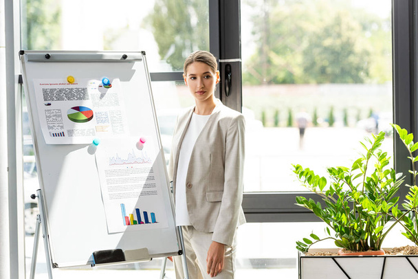 Selbstbewusste Geschäftsfrau in formaler Kleidung steht neben Flipchart mit Infografik im Büro - Foto, Bild