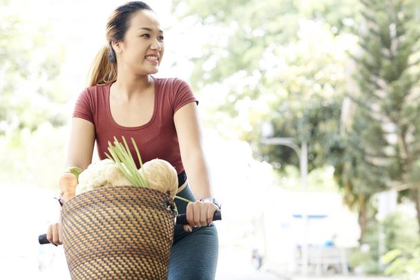 Happy young Vietnamese woman riding home from local market with basket full of fresh vegetables - Photo, Image