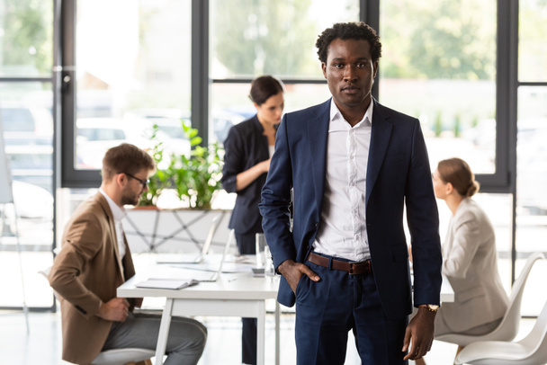 オフィスでポケットに手を入れて立っているフォーマルな服装でアフリカ系アメリカ人ビジネスマンの正面図 - 写真・画像