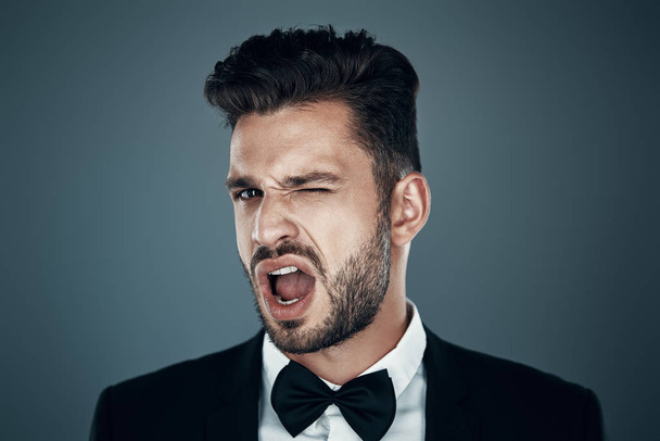 Charming young man in bow tie looking at camera and making a face while standing against grey background - Fotoğraf, Görsel
