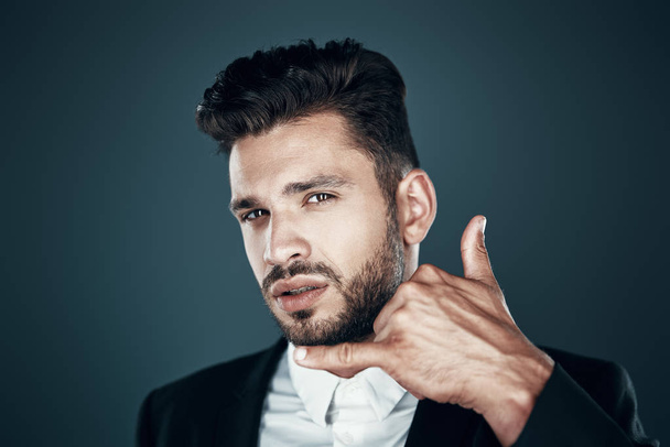 Charming young man in formalwear showing call me gesture and looking at camera while standing against grey background - Photo, Image