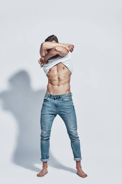 Full length of handsome young man taking off his T-shirt while standing against grey background - Fotografie, Obrázek