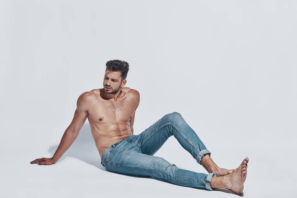 Handsome young shirtless man looking away while sitting against grey background - Zdjęcie, obraz