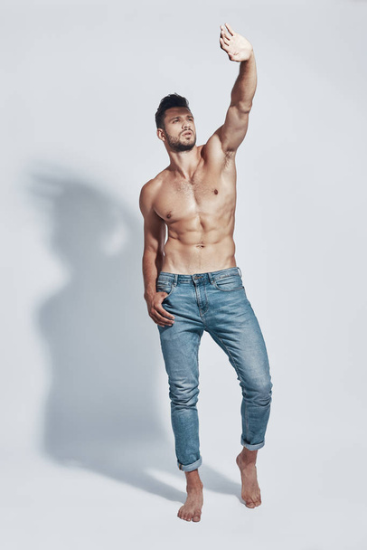 Full length of handsome young shirtless man covering from light with hand while standing against grey background - Valokuva, kuva