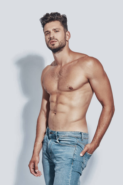 Handsome young shirtless man looking at camera while standing against grey background - Foto, imagen