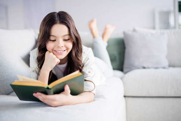 Portrait of her she nice attractive lovely charming cute cheerful cheery focused wavy-haired preteen girl lying on divan reading novel in light white interior living-room indoors - Foto, Imagen