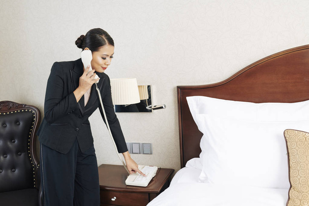 Asian młody biznesmen w formalwear trzyma słuchawkę i ma zamiar zadzwonić na recepcji do obsługi pokoju, stojąc w pokoju hotelowym - Zdjęcie, obraz