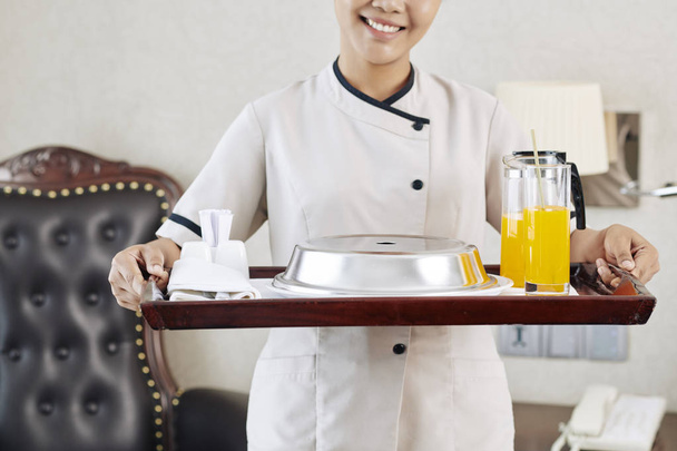 Nahaufnahme einer jungen Kellnerin in Uniform, die ein Tablett mit Frühstück und Saft in der Hand hält und im Hotelzimmer lächelt - Foto, Bild