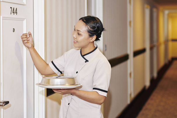 Asiática joven camarera en uniforme blanco celebración de la cena en la bandeja y llamando a la puerta, mientras que de pie en el pasillo del hotel
 - Foto, Imagen