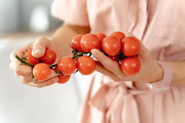 Woman holding twig of ripe cherry tomatoes in the kitchen. Concept of healthy eating and dieting lifestyle - 写真・画像