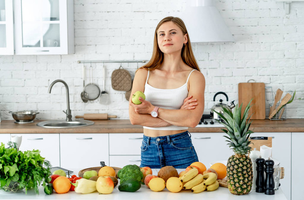 Young woman standing near desk in the kitchen, smiling, looking at camera. Healthy food and Dieting concept. - Foto, imagen