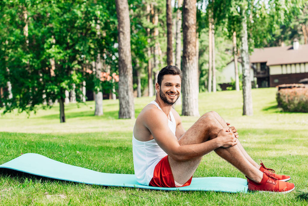Fröhlicher bärtiger Sportler sitzt auf Fitnessmatte im Park  - Foto, Bild