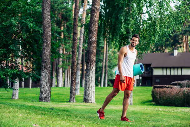 cheerful bearded man holding fitness mat while walking in park  - Foto, afbeelding