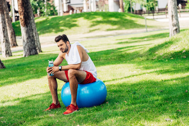bearded sportsman holding sport bottle while sitting on fitness ball - Photo, Image