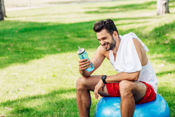 glücklicher bärtiger Mann schaut auf Sportflasche, während er auf Fitnessball sitzt  - Foto, Bild