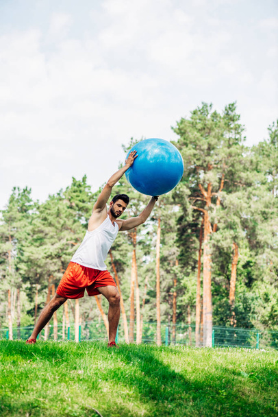 όμορφος μουσάτος αθλητής δουλεύοντας έξω κρατώντας μπάλα γυμναστικής στο πάρκο  - Φωτογραφία, εικόνα
