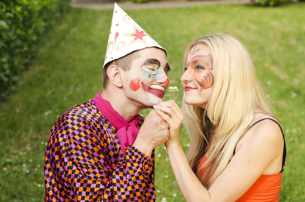 Porträt eines lächelnden Mannes, der wie ein Clown gekleidet versucht, einem glücklichen Mädchen mit Schmetterling-Make-up eine Blume zu überreichen - Foto, Bild