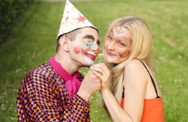 Портрет улыбающегося человека, одетого как клоун, пытающегося подарить цветок счастливой девушке с макияжем бабочки
 - Фото, изображение