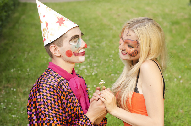 Porträt eines lächelnden Mannes, der wie ein Clown gekleidet versucht, einem glücklichen Mädchen mit Schmetterling-Make-up eine Blume zu überreichen - Foto, Bild