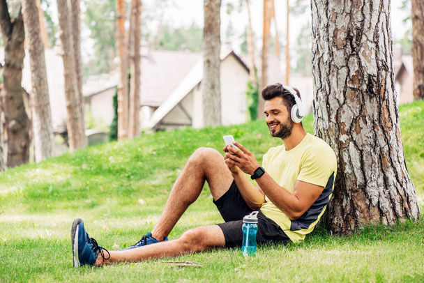 veselý muž ve sluchátkách, používající smartphone blízko kmene stromů a poslech hudby  - Fotografie, Obrázek