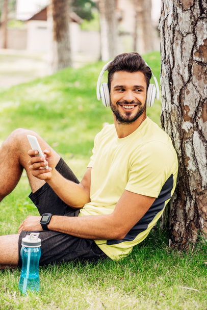 positive man in headphones using smartphone near tree trunk and listening music  - Photo, Image