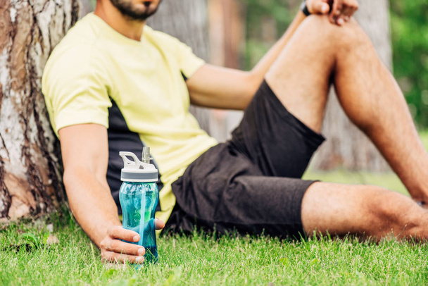 Ausgeschnittene Ansicht eines bärtigen Sportlers mit Sportflasche in der Nähe eines Baumstammes  - Foto, Bild