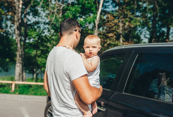 Vader in shirt staan in de buurt van auto met zijn baby. Klein meisje met nieuwsgierigheid op zoek in de camera. - Foto, afbeelding