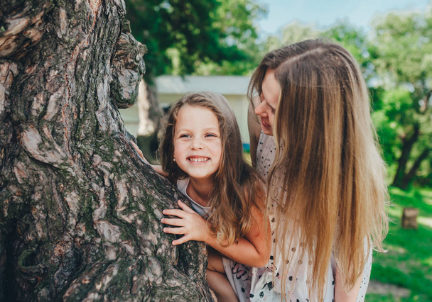 Mãe jovem e filhinha fofa se divertindo perto de uma árvore grande em um parque de verão
. - Foto, Imagem