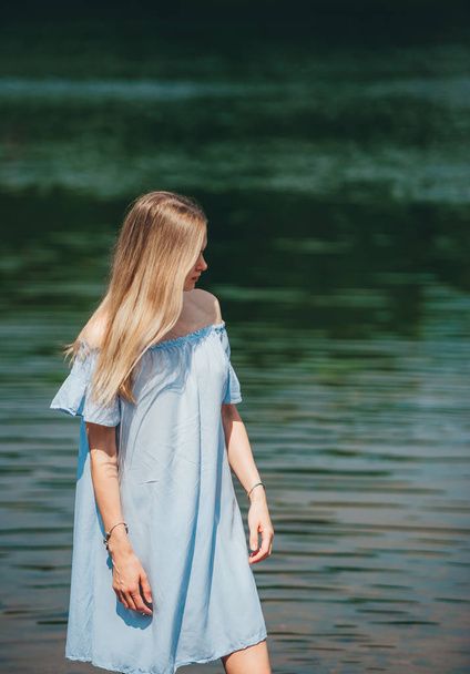 Portré a szép fiatal nő, hosszú hajú visel kék ruha állt, közel a tó partján a napsütéses nyári napon. - Fotó, kép