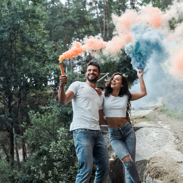 attractive woman and cheerful bearded man holding smoke bombs outside  - Zdjęcie, obraz