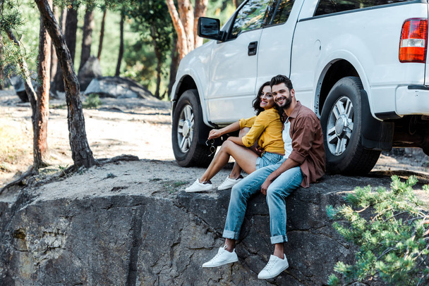 gelukkige bebaarde man en aantrekkelijke vrouw zitten in de buurt van auto en bomen  - Foto, afbeelding