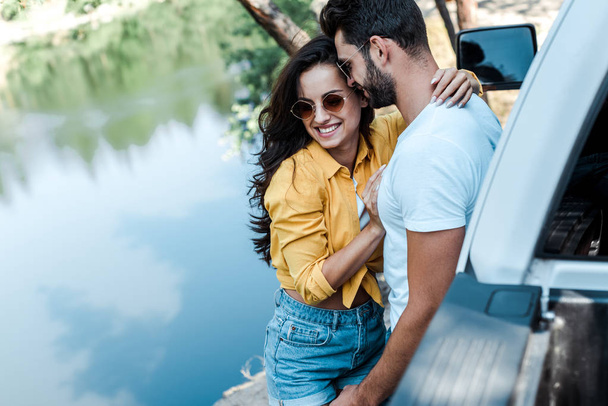 selective focus of happy girl hugging with boyfriend near car and river  - Foto, Bild