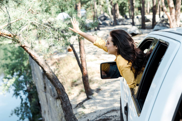 cheerful girl waving hand from car window in woods  - Photo, Image