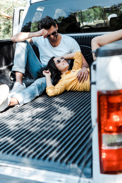 selective focus of bearded man in sunglasses sitting in car trunk and looking at girl  - Фото, изображение