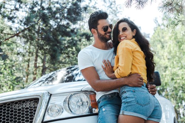 low angle view of man standing and hugging happy girl near auto in woods  - Foto, imagen