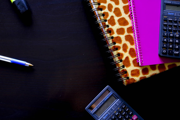 Composition d'objets de bureau composés de deux carnets avec deux calculatrices, un stylo bleu et un surligneur sur un fond noir vu d'en haut
 - Photo, image