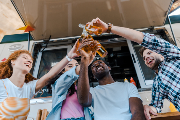 Tiefblick auf fröhliche multikulturelle Freunde, die in der Nähe von Foodtrucks Flaschen mit Bier klappern   - Foto, Bild