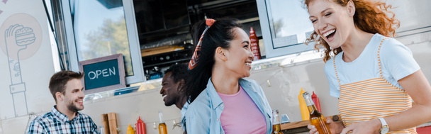 panoramische foto van meisjes lachen in de buurt van multiculturele mannen en voedsel vrachtwagen  - Foto, afbeelding