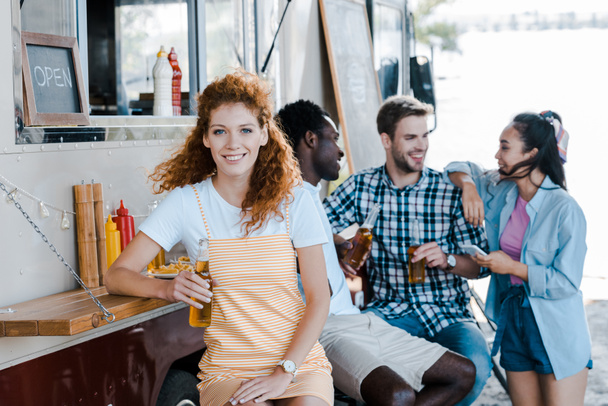 enfoque selectivo de chica pelirroja feliz sosteniendo botella con cerveza cerca de amigos y camión de comida
  - Foto, Imagen