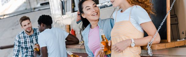 panoramische foto van gelukkige meisjes die flessen met bier in de buurt van multiculturele mannen en Food truck houden  - Foto, afbeelding