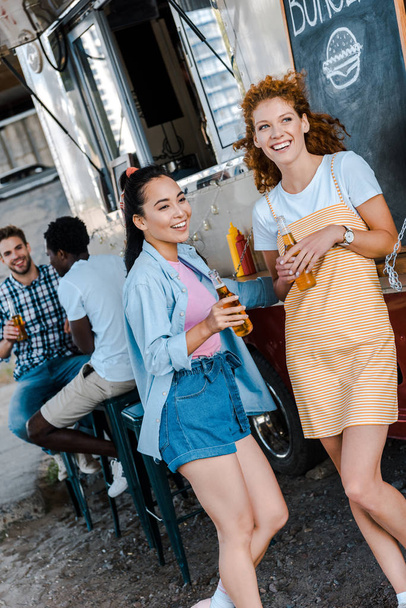 selectieve focus van gelukkige vrouwen die flessen met bier in de buurt van multiculturele mannen en Food truck houden  - Foto, afbeelding