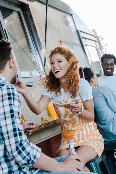 Selektiver Fokus des rothaarigen Mädchens, das lacht, während es Mann in der Nähe multikultureller Menschen füttert  - Foto, Bild