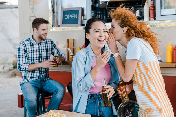selective focus of redhead girl whispering in ear of happy woman laughing near multicultural men  - 写真・画像