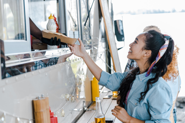 bijgesneden beeld van Afro-Amerikaanse man geven kartonnen plaat aan Aziatische meisje in de buurt van Food truck  - Foto, afbeelding